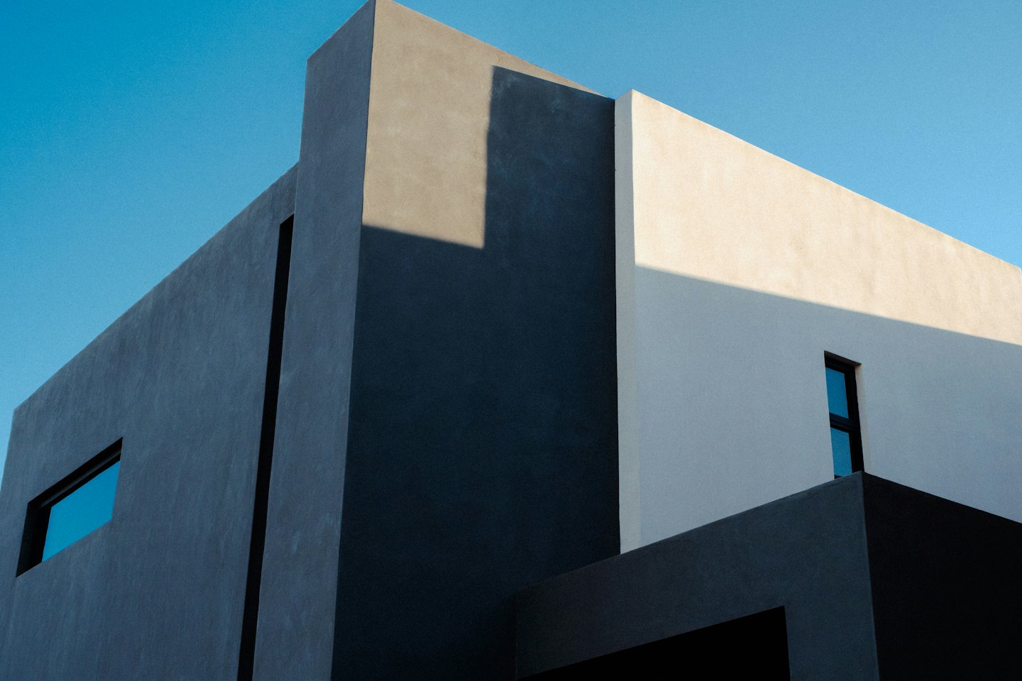 Moderná betónová budova s geometrickými tvarmi a oknami na pozadí modrej oblohy.